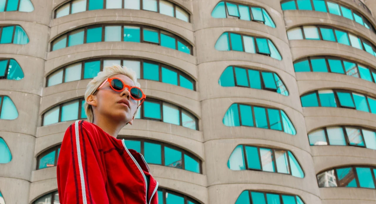Femme en veste rouge portant des lunettes de soleil Theo Eyewear rouges devant un immeuble aux vitres turquoise